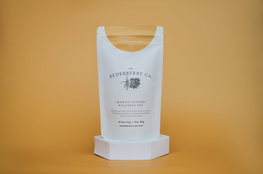 The Elderberry Co. Immune Support Wellness Tea, Lemon Ginger loose leaf herbal tea pouch on a white display stand against a warm yellow background.