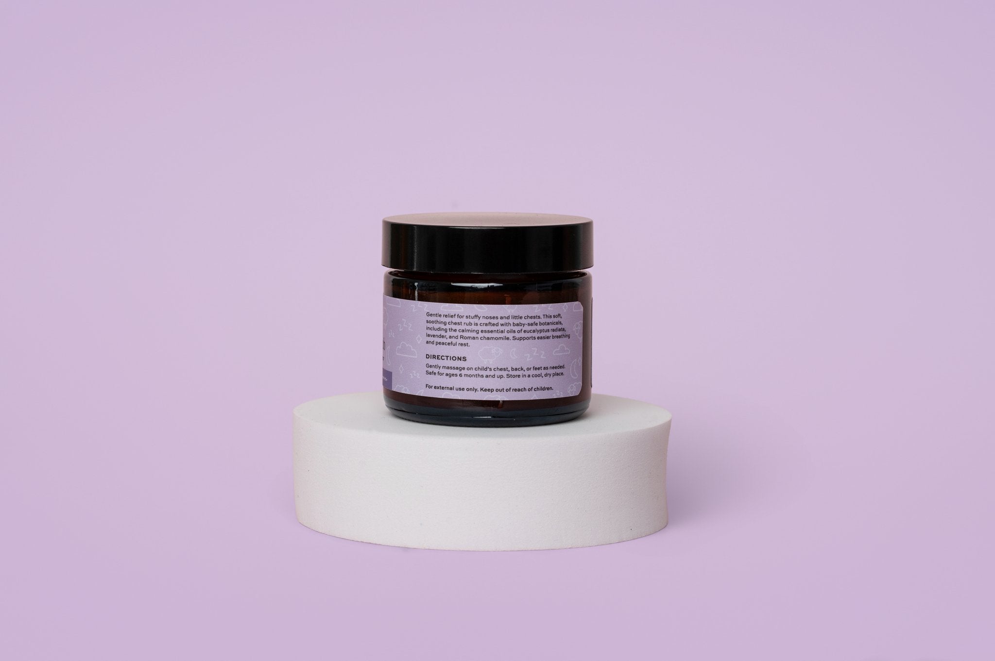 Back view of The Elderberry Co. Baby & Kids Chest Rub jar displaying usage directions and ingredients on a white pedestal against a soft lavender background.