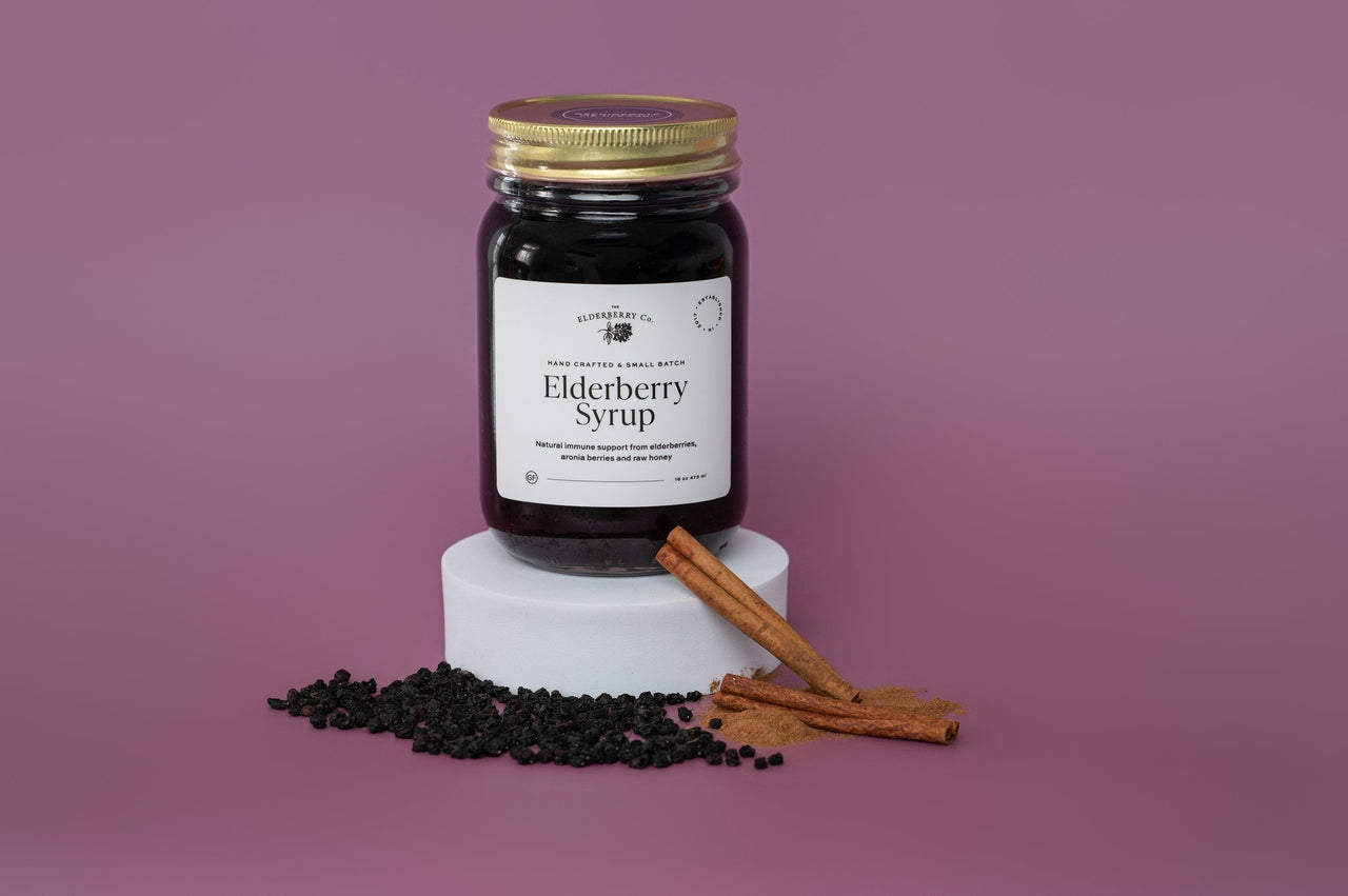 Jar of Elderberry Syrup on a white pedestal with cinnamon sticks and elderberries against a purple background