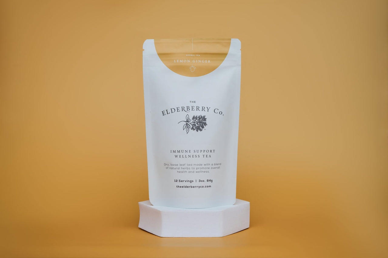 The Elderberry Co. Immune Support Wellness Tea, Lemon Ginger loose leaf herbal tea pouch on a white display stand against a warm yellow background.