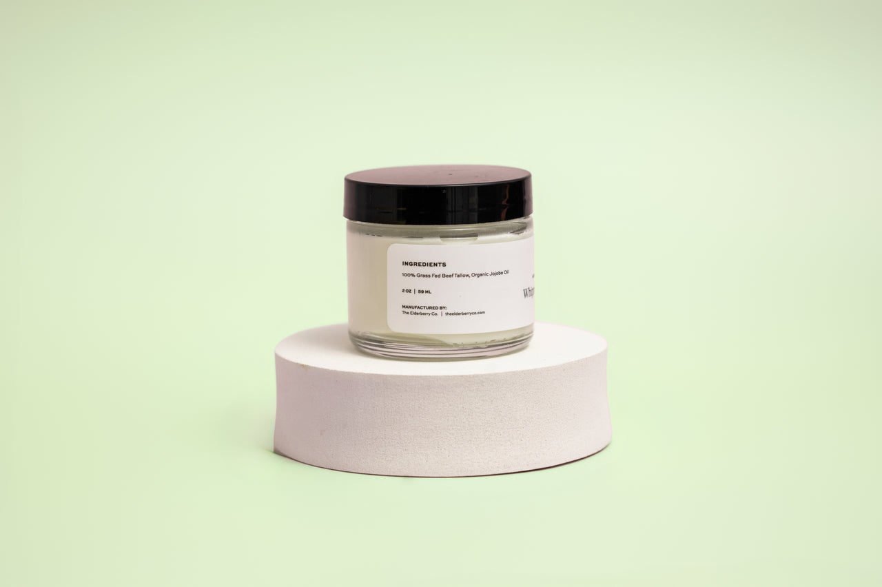 Back view of The Elderberry Co. Nourishing & Hydrating Tallow Balm jar displayed on a white pedestal against a soft light green background, showing the ingredient panel and product information label.
