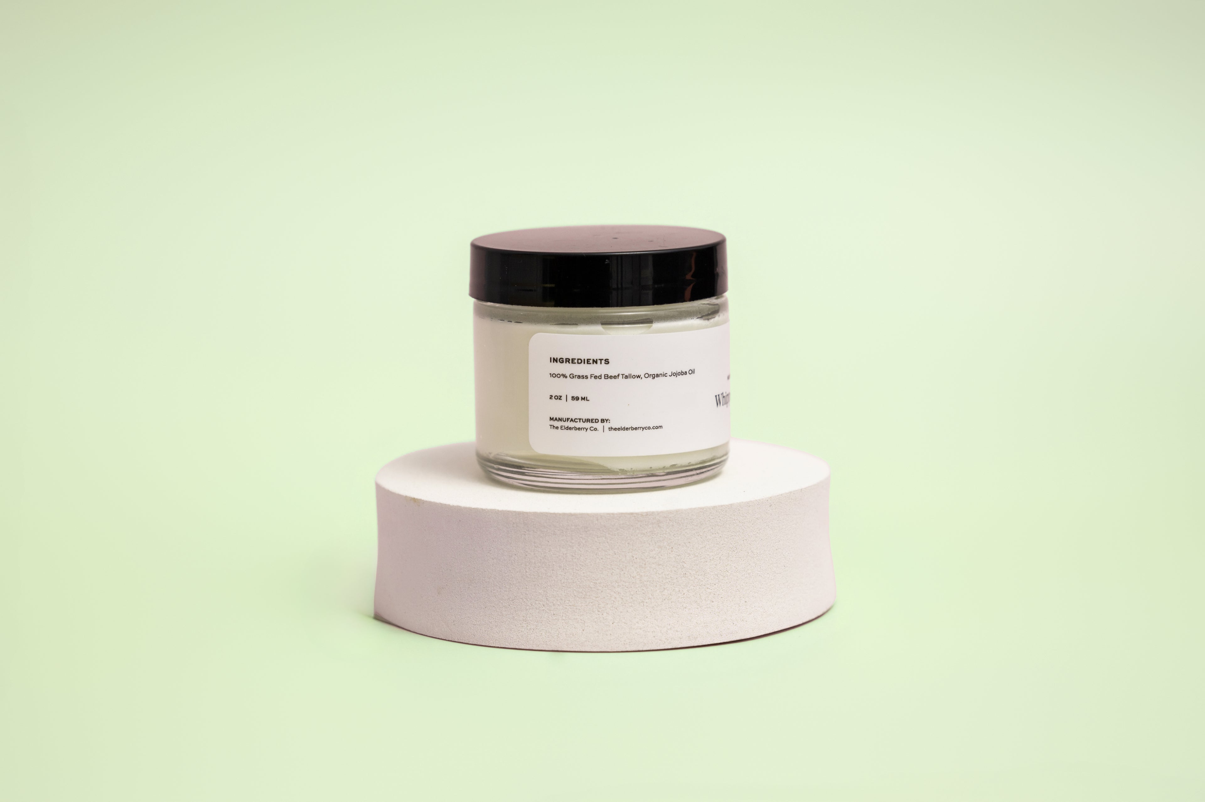 Back view of The Elderberry Co. Nourishing & Hydrating Tallow Balm jar displayed on a white pedestal against a soft light green background, showing the ingredient panel and product information label.
