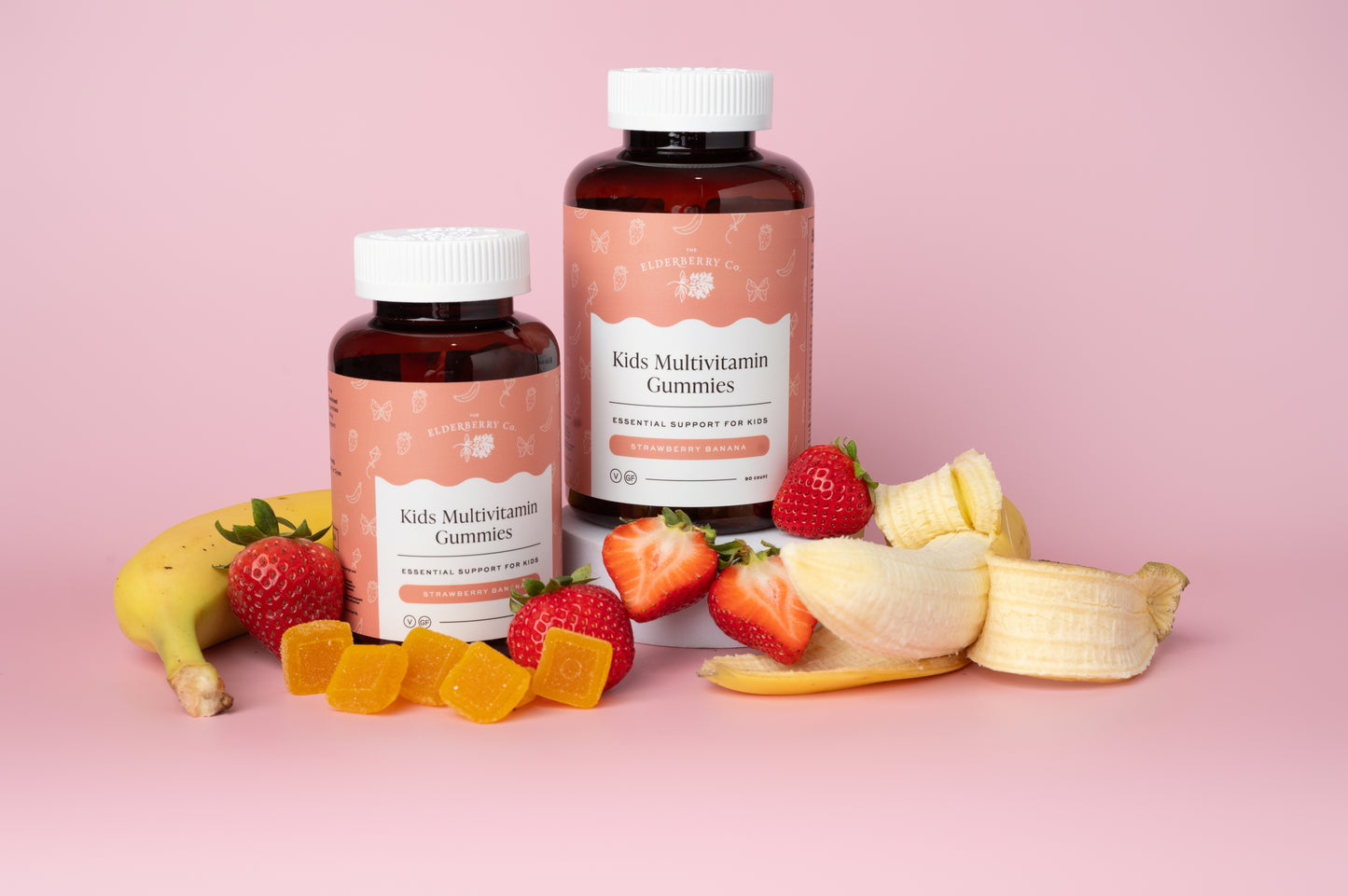 Two bottles of Kids Multivitamin Gummies with gummy bears, strawberries, and bananas on a pink background.