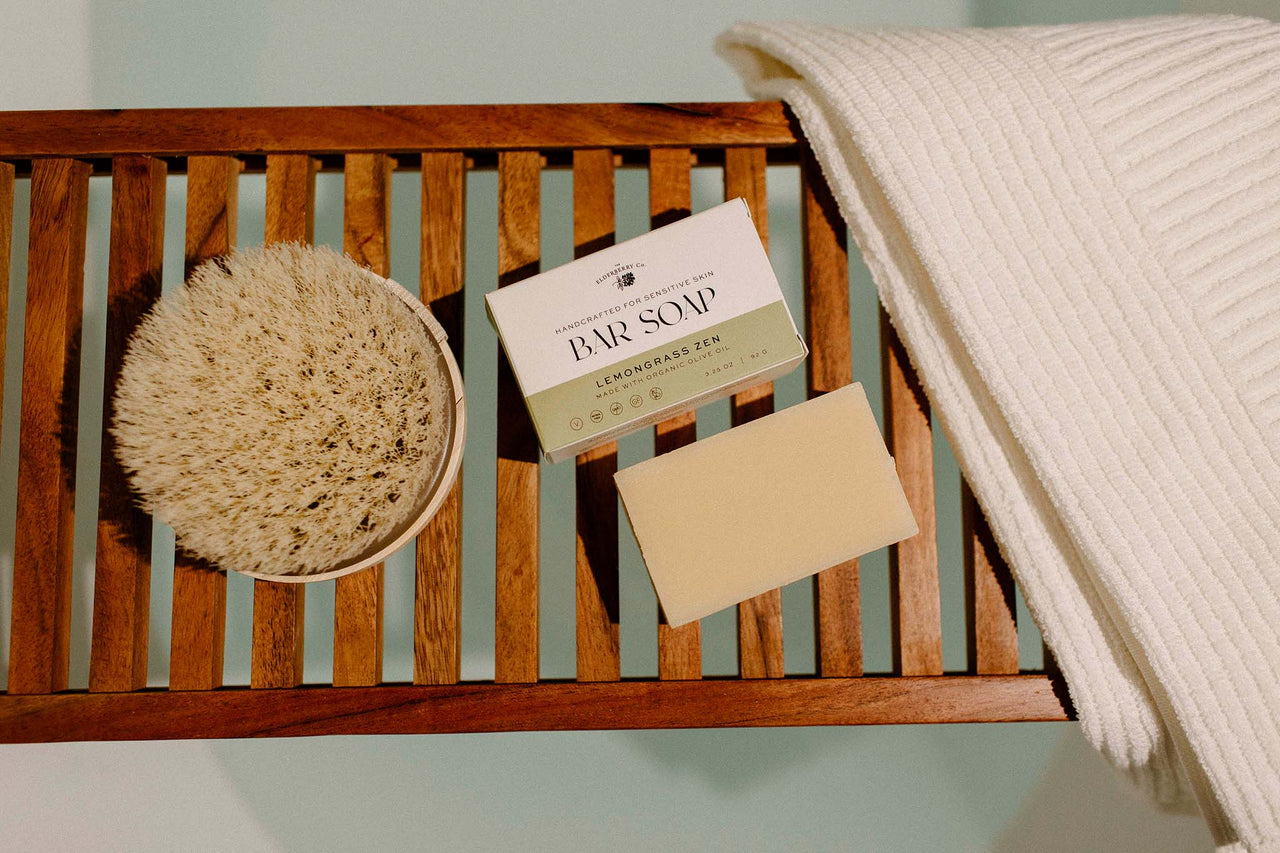 The Elderberry Co. Lemongrass Zen bar soap with packaging, natural exfoliating brush, and towel arranged on a wooden bath bench.
