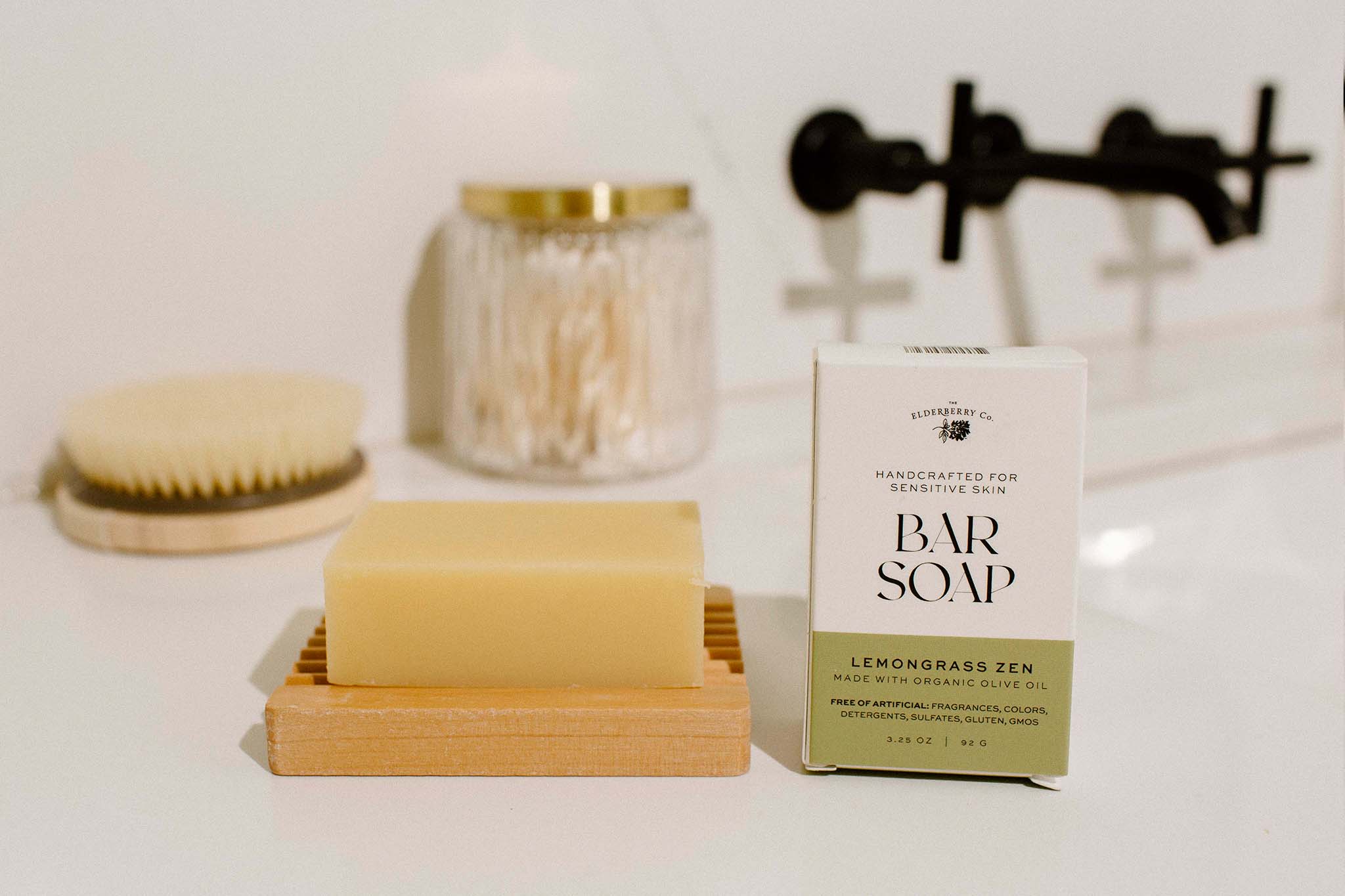 The Elderberry Co. Lemongrass Zen bar soap and packaging displayed on a wooden soap dish beside a bathtub with natural bath accessories.