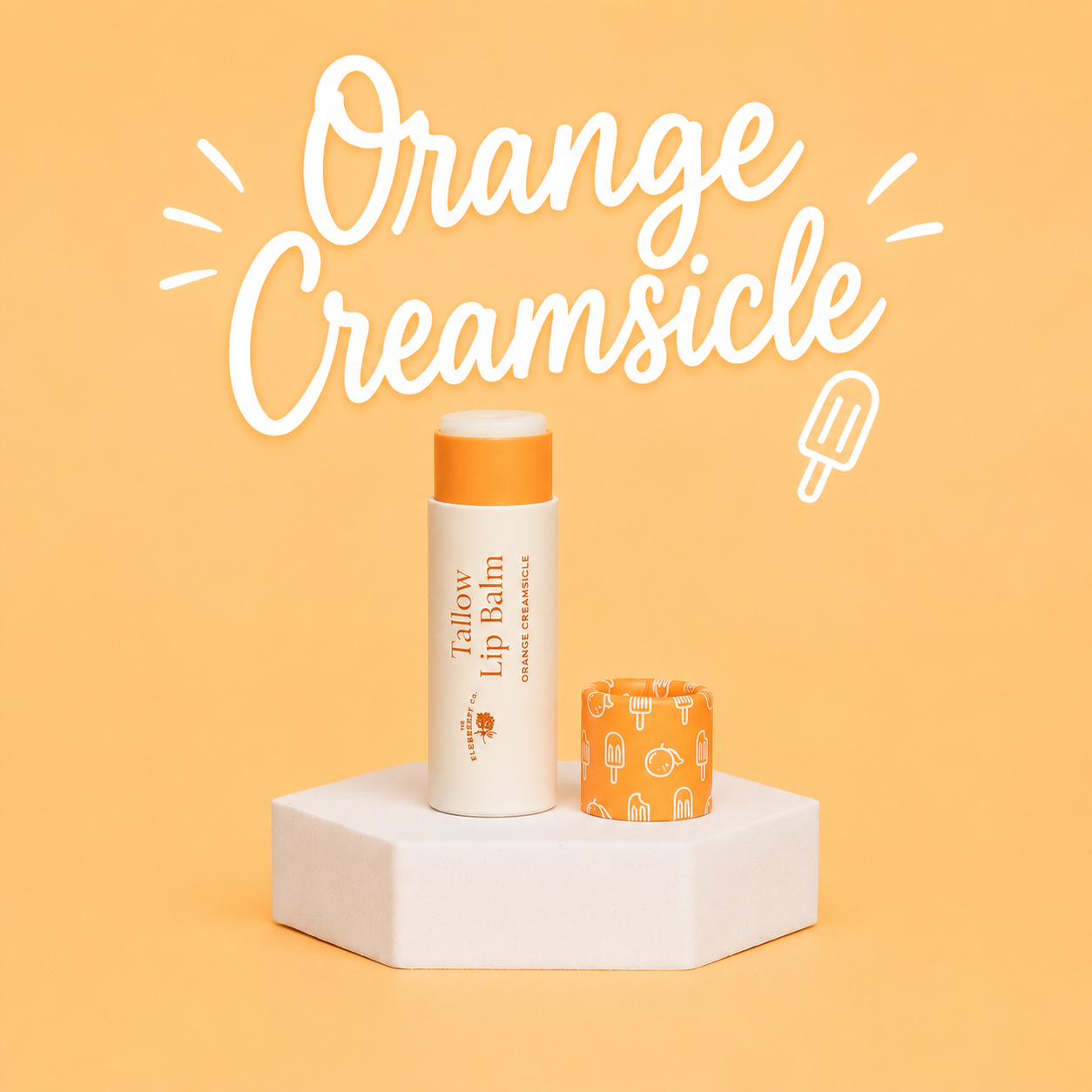 A tube of Farrow & Ball Orange Creamsicle lip balm with its cap off, displayed on a white hexagonal pedestal against a warm orange background with white hand-drawn text reading 'Orange Creamsicle'.