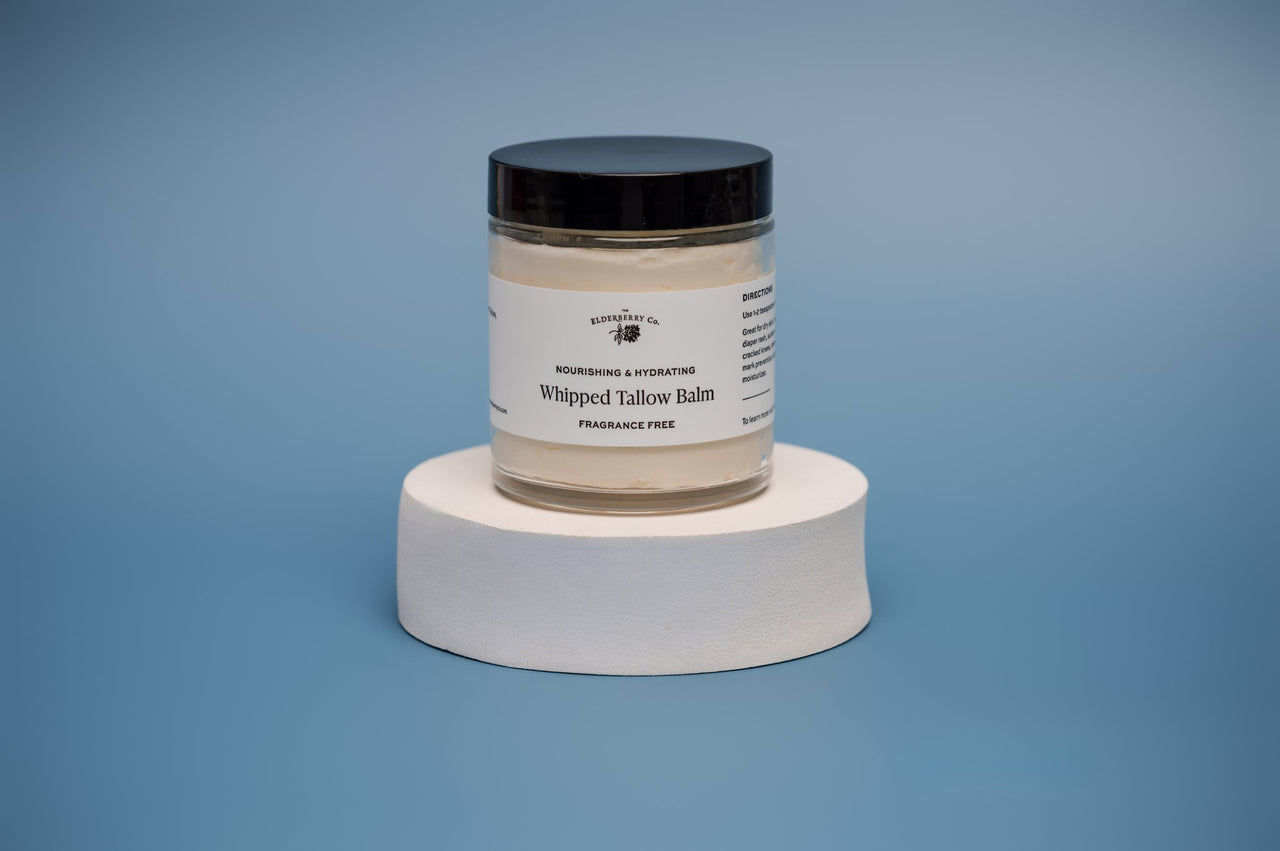 Three jars of Fragrance Free Whipped Tallow Balm, with one jar open showing the creamy product inside. The jars are placed on a white surface with a blue background.