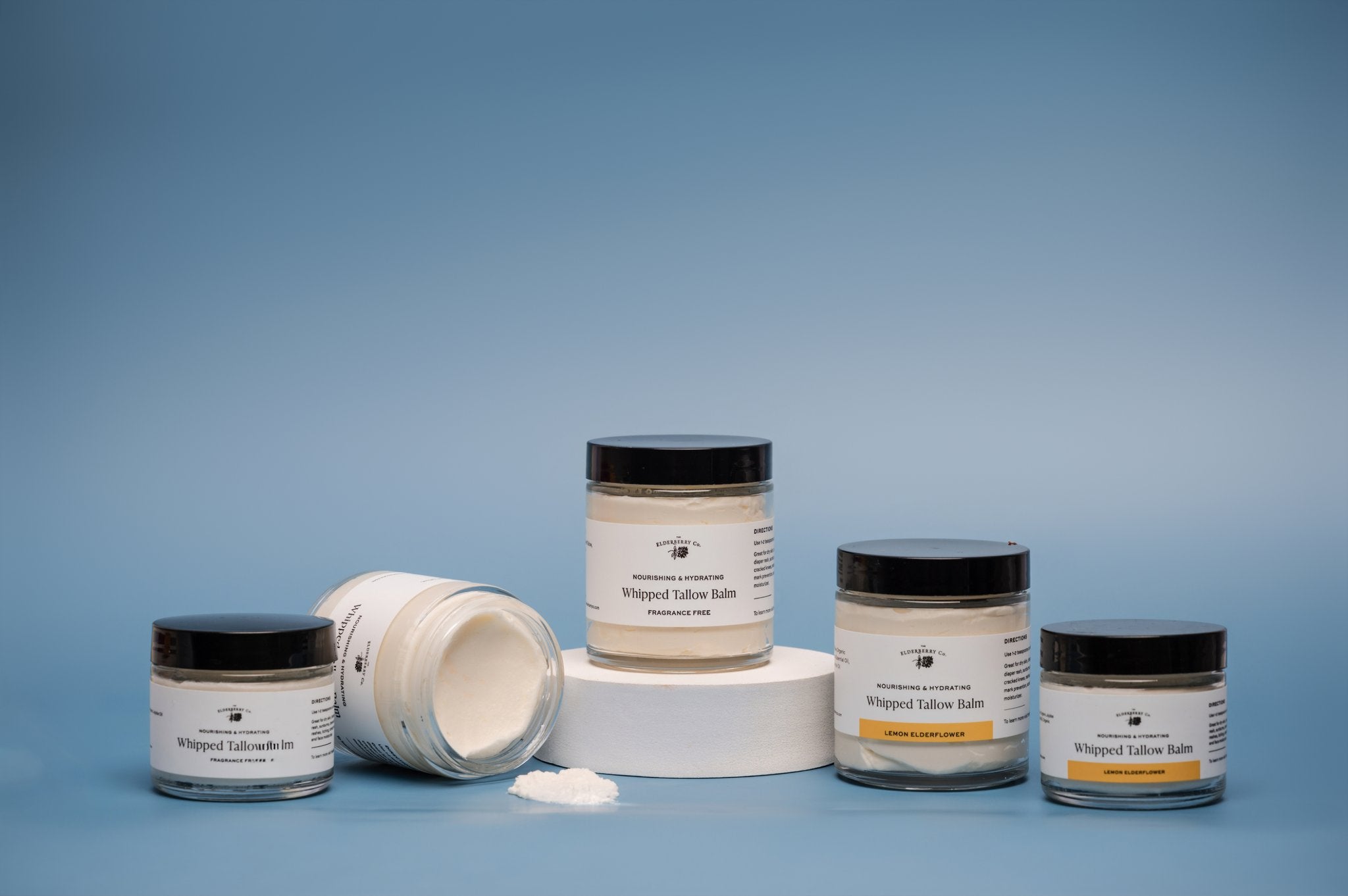 Collection of The Elderberry Co. Whipped Tallow Balm jars in Fragrance Free and Lemon Elderflower varieties displayed on a blue background, including open and closed jars showcasing the whipped balm texture.