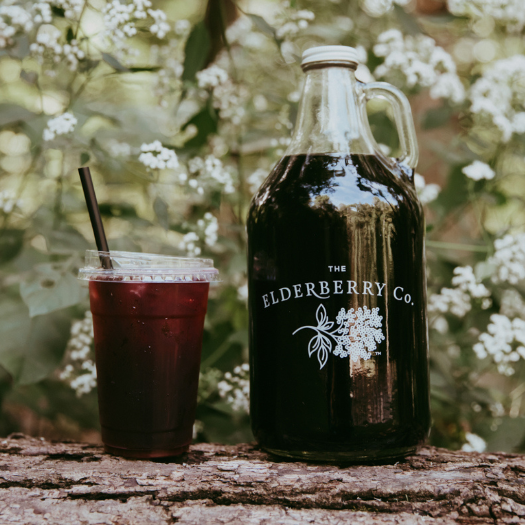 The Elderberry Co. elderberry syrup growler and iced elderberry drink displayed outdoors on a log with a forest backdrop.